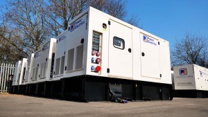 A row of large, white Power Electrics generators are lined up outdoors on a paved surface, connected and ready for use, with trees and a metal fence visible in the background; clear blue sky overhead suggests a sunny day.